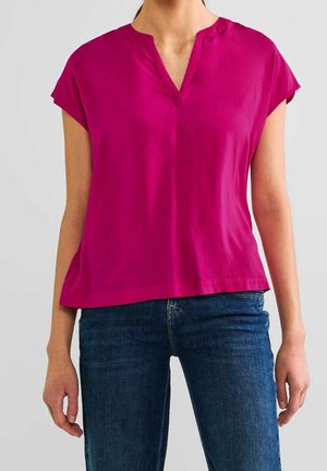 Femme portant un chemisier magenta à manches courtes et col en V, associé à un jean bleu foncé sur un fond clair uni.