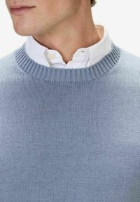 Ljusblå stickad tröja med ribbad rund hals, lagd över en vit skjorta med krage. Slät textur och jämn sömnadsdetalj synlig.