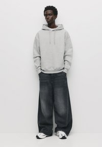 Felpa grigia chiara con cappuccio e stringhe, dotata di tasca frontale, abbinata a jeans scuri a taglio ampio. Le scarpe sono sneakers sportive bianche.