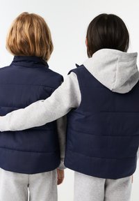 Gilet imbottiti blu navy con colli alti, abbinati a felpe grigie con cappuccio. I gilet presentano pannelli imbottiti orizzontali e tasche laterali.
