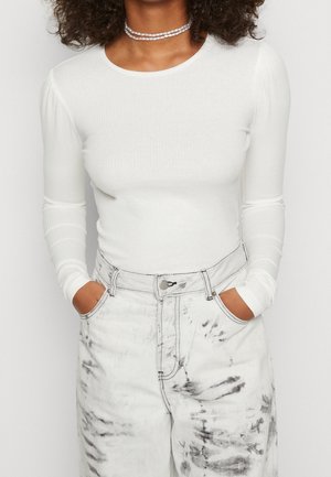 Wit geribd langärmelig shirt gecombineerd met een lichtgekleurde high-waist denim jeans met een gemarmerd patroon, met handen in de zakken.