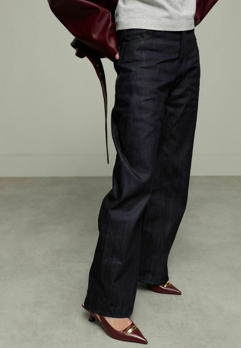 Pantalons en denim bleu foncé à jambes larges avec une texture lisse, associés à des talons aiguilles bordeaux dotés d'accents dorés.