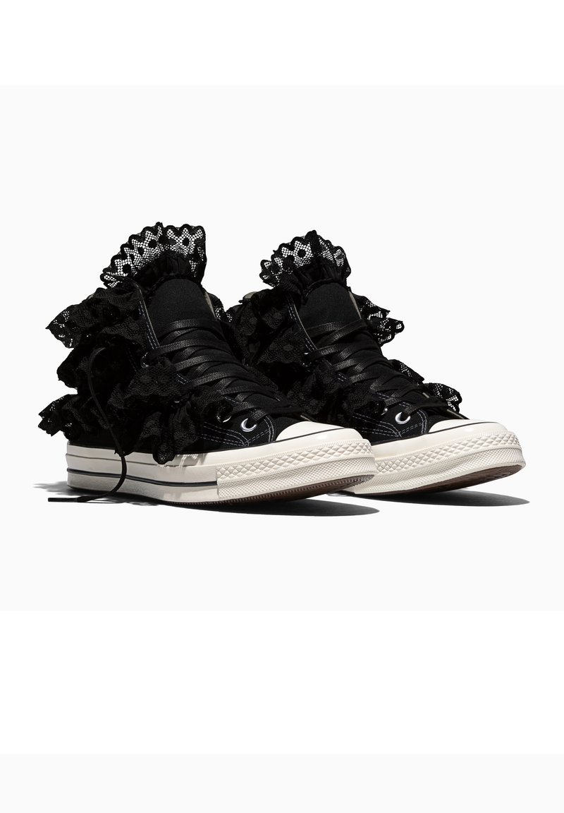 Zapatillas negras de caña alta con suelas blancas, con volantes de encaje negro en capas alrededor de los tobillos y cordones negros.