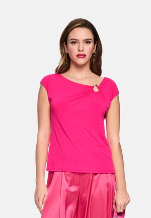 Frau mit langen braunen Haaren, die ein knallpinkes kurzärmeliges Oberteil mit Ringdetail an einer Schulter und einen passenden pinken Satinsrock trägt.