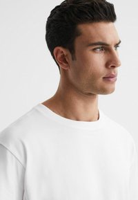 T-shirt bianco in cotone con scollo rotondo, vestibilità rilassata e texture liscia, mostrato sul corpo superiore e sul profilo di un modello contro uno sfondo neutro.