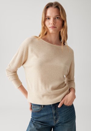 Pull en tricot beige avec des manches trois quarts, un col rond et un motif texturé. Associé à un jeans en denim bleu.