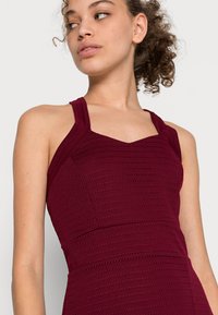 Vestido sem mangas de cor bordô com um design texturizado em ribetes. Apresenta um corte justo e um decote em V com alças largas e costuras limpas.