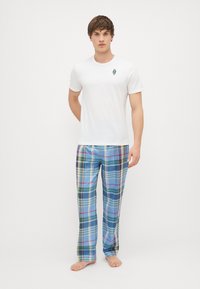 Balta kokvilnas T-kreklā ar mazu zilu un zaļu emblēmu krūšu rajonā, apvienota ar daudzkrāsainiem plāna raksta pidžamu biksēm zilo, zaļu un rozā toņu kombinācijā.