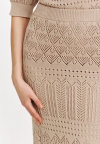Beżowa sukienka dzianinowa z misternym wzorem; posiada teksturowane szycie, otwarte sekcje oraz prążkowany pas.