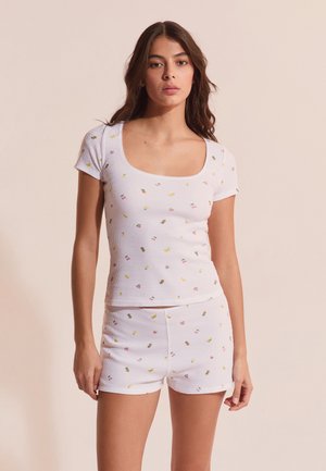 Młoda kobieta ubrana w biały, prążkowany top z krótkim rękawem i spodenki z małymi, kolorowymi wzorami owoców, stojąca na beżowym tle.