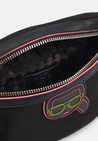 Svart dragkedjedosa med rosa detaljer, grafisk bild av neongula glasögon och mönstrad svart tygfoder med varumärkesetikett.