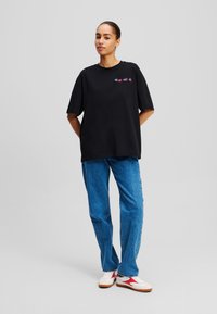 T-shirt en coton noir avec une coupe oversize, manches courtes, et un graphique coloré brodé sur la poitrine, associé à un jean droit bleu et des baskets blanches.