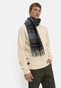 Mann in beigem Pullover und dunklen Hosen mit einem blau-braun karierten Schal um den Hals, stehend vor einem schlichten Hintergrund.