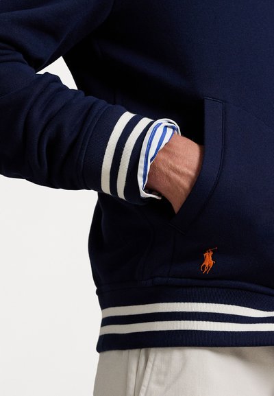 Námornícky sveter s bielymi pruhovanými manžetami a malým oranžovým logom polo. Hladká tkanina, voľný strih a detail predné vrecko.