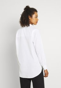 Camicia bianca a maniche lunghe con orlo arrotondato, polsini con bottone e texture liscia. Design su misura con dettaglio della cucitura posteriore.