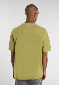 Muž s tetováním na krku a rukou, oblečený v jednoduchém olivově zeleném tričku s krátkým rukávem a šedých kalhotách, stojící zády k bílé stěně.