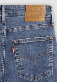 Jean en denim bleu avec un patch en cuir marron à la ceinture. Comprend des coutures classiques sur les poches arrière et une petite étiquette rouge Levi's.