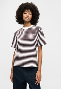 Νεαρό άτομο με κατσαρά μαλλιά που φοράει λευκό και σκούρο ριγέ κοντομάνικο πουκάμισο και σκούρο παντελόνι, στέκεται μπροστά σε απλό φόντο.