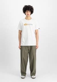 Balta medvilninė marškinėliai su auksiniu ir juodu "ALPHA INDUSTRIES" logo, derinami su žaliais, laisvais keliais ir baltais sportbačiais.