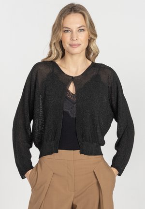 Czarny, przejrzysty, dzianinowy kardigan z długimi rękawami, dekoltem w serek i zapięciem na guziki z przodu, w połączeniu z czarną koronkową bluzką i brązowymi spodniami.