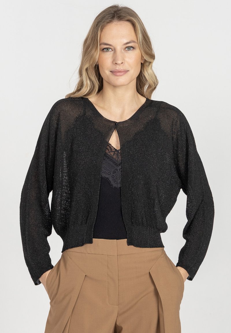 Czarny, przejrzysty, dzianinowy kardigan z długimi rękawami, dekoltem w serek i zapięciem na guziki z przodu, w połączeniu z czarną koronkową bluzką i brązowymi spodniami.