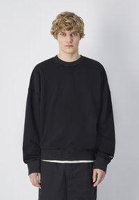 Sudadera negra con cuello redondo, hombros caídos y puños acanalados. La tela parece suave con una ligera textura. Diseño minimalista.