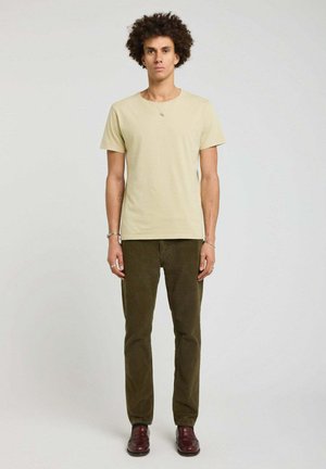 Jeune homme debout face à l'avant portant un t-shirt beige, un pantalon vert olive et des chaussures rouge foncé sur un fond clair uni.