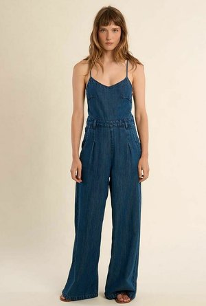 Donna con capelli castano chiaro che indossa una tuta blu in denim senza maniche con gambe larghe e spalline sottili, in piedi davanti a uno sfondo beige uniforme.