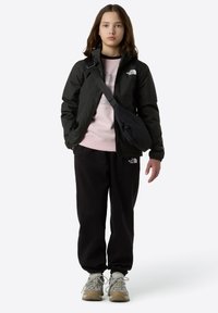 Veste noire imperméable, sweat-shirt rose clair, pantalon noir, baskets grises avec accents jaunes et sac banane noir. Tenue décontractée d'extérieur.