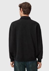 Mand med kort brunt hår iført en sort langærmet skjorte og blå jeans, stående med ryggen til mod en ensfarvet baggrund.