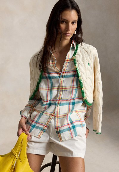 Ung kvinne iført en kremfarget kabelstrikket cardigan over en rutete skjorte og hvite shorts, holder en knallgul håndveske.