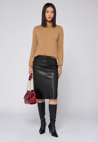 Braunfarbener Langarmoberteil, schwarze Bleistift-Rock aus Leder, schwarze kniehohe Stiefel. Das Model hält eine rote Handtasche mit goldenen Akzenten.