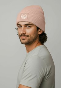 Pink ribbed knit beanie with a logo patch featuring waves, worn with a light gray t-shirt. The fabric appears soft with a textured finish.