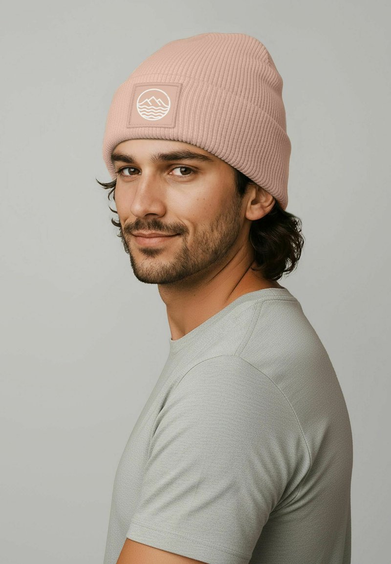 Pink ribbed knit beanie with a logo patch featuring waves, worn with a light gray t-shirt. The fabric appears soft with a textured finish.