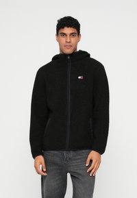 Musta fleece-omalla vetoketjulla varustettu huppari, jossa on pieni logo rinnassa ja sivutaskut, teksturoitu kangas ja rento malli.