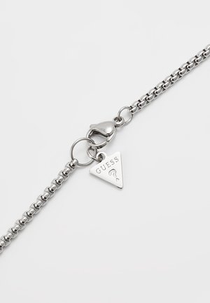 Zilveren kettingarmband met een karabijnsluiting en een kleine driehoekige hanger gegraveerd met het woord "GUESS" en een vraagteken.