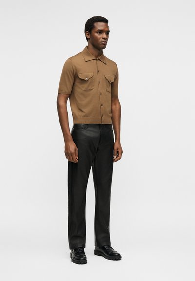 Άνδρας που στέκεται, φορά καφέ κοντομάνικο πουκάμισο με τσέπες, μαύρο δερμάτινο παντελόνι και μαύρα παπούτσια σε λευκό φόντο.