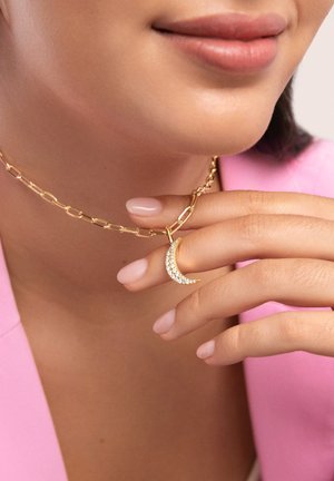 Goldene Kettenhalskette mit verlängerten Gliedern und einem Anhänger in Form eines Halbmonds, der mit klaren Strasssteinen verziert ist. Sanfter rosafarbener Hintergrund, neutrale Nagellackfarbe.