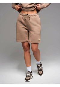 Pantaloni corti in cotone beige con cintura elastica e cordoncino, dotati di tasche laterali e un'etichetta sull'orlo. Abbinate a sneakers marroni e calzini bianchi.