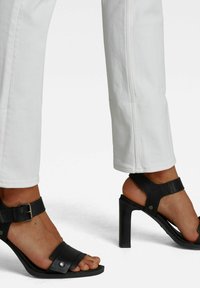 Sandalias de tacón alto negras con detalles de correas y herrajes plateados, combinadas con pantalones blancos cortos. Muestra una textura de cuero suave.