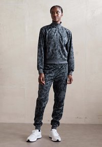 Grå leopardmønstrede treningsdresser laget av mykt stoff, med jakke med glidelås og bukser med elastisk midjebånd; satt sammen med hvite sneakers.