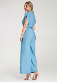 Niebieski kombinezon z kołnierzykiem, krótkimi rękawami i wiązaniem w talii. Materiał wydaje się lekki o gładkiej fakturze. Stylizowany z przezroczystymi obcasami.
