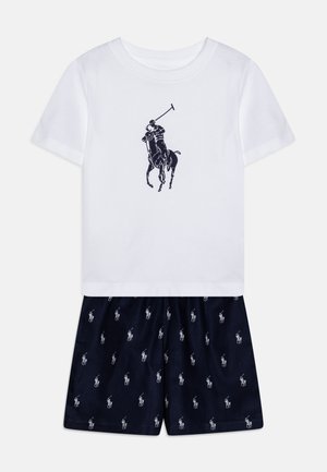 Biela bavlnená tričko s logom námorníckeho polo hráča, v kombinácii s námorníckymi šortkami, ktoré majú opakovaný malý biely vzor polo hráča.