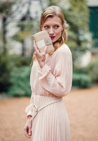 Žena v bledě růžových šatech drží béžovou pen�ěženku blízko své tváře, hledí do strany venku s rozostřenou zelení v pozadí.