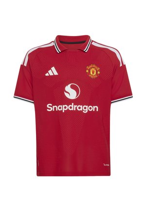 Chłopiec w stroju piłkarskim Manchester United, trzymający czarno-białą piłkę Adidas, stoi na jednolitym tle z cieniem.