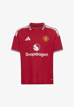 Niño con equipación del Manchester United sosteniendo una pelota Adidas negra y blanca, de pie sobre un fondo liso con sombra proyectada.
