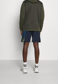 Sweat à capuche vert foncé avec un design texturé, assorti à un short bleu marine présentant un motif dégradé en vert et bleu sur le côté.