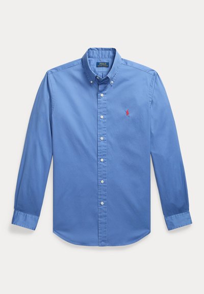 Chemise bleue à manches longues avec col boutonné, boutons blancs et petit logo rouge brodé sur la poitrine gauche.