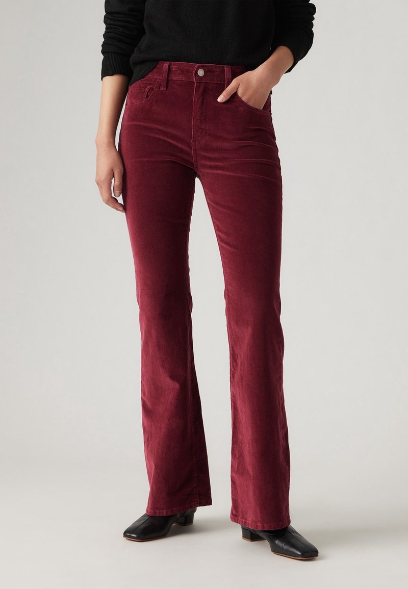 Personne portant un pantalon flare en velours côtelé bordeaux, des bottines noires à bout carré, et un haut noir à manches longues, une main dans la poche.