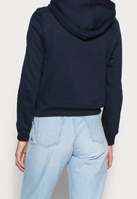 Felpa corta blu navy con cappuccio e orlo a costine, abbinata a jeans leggeri a vita alta con cuciture delicate sulle tasche posteriori.